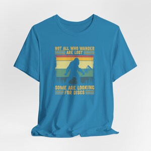 May include: A blue t-shirt with a vintage-style graphic of a Bigfoot holding a frisbee. The graphic says "Not all who wander are lost. Some are looking for discs."
