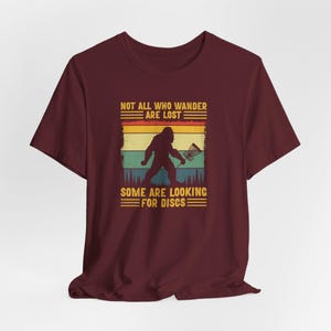 May include: A maroon t-shirt with a vintage-style graphic of a Bigfoot holding a disc golf disc. The text reads "Not all who wander are lost. Some are looking for discs."