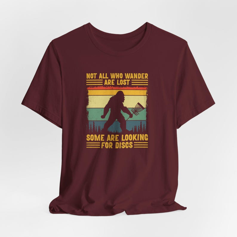 May include: A maroon t-shirt with a vintage-style graphic of a Bigfoot holding a disc golf disc. The text reads "Not all who wander are lost. Some are looking for discs."