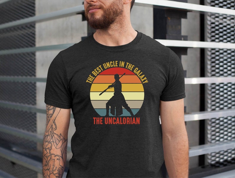 May include: A dark grey t-shirt with a vintage-style graphic of a person holding a lightsaber. The graphic says "The Best Uncle in the Galaxy The Uncalorian".