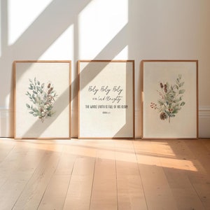 May include: Three framed art prints with botanical illustrations and text. The prints feature watercolor images of leaves, flowers, and pinecones. One print displays the text "Holy Holy Holy is the Lord Almighty" from Isaiah 6:3. The frames are a light brown colour.