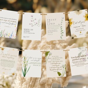 May include: A collection of eight rectangular cards with inspirational quotes and floral designs, strung on a twine. The cards feature various colors, including white, green, yellow, and purple, with text from the Bible. The background is a blurred, natural setting.