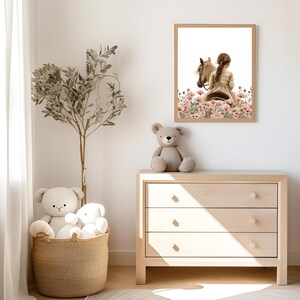May include: A framed illustration of a girl riding a horse through a field of pink flowers. The illustration is in a light brown frame and is hanging on a white wall. There are two stuffed animals in the foreground.