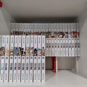 Può includere: Un set completo di volumi manga di One Piece, numerati dal 27 al 38, con il nome dell'autore, Eiichiro Oda, stampato sui dorsi. I volumi sono disposti su uno scaffale bianco.