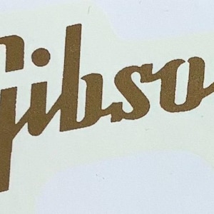 Pode incluir: Logotipo "Gibson" dourado sobre fundo creme. O logotipo está em itálico, com as letras do nome da marca conectadas. O adesivo tem um formato arredondado e irregular.
