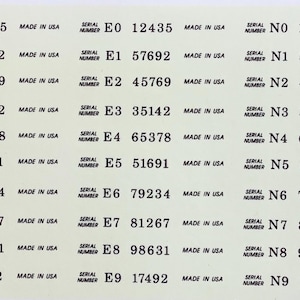 Op de afbeelding: Een vel witte etiketten met zwarte tekst. Elk etiket toont een serienummer en "MADE IN USA". De etiketten zijn gerangschikt in rijen en kolommen, waarbij elke rij drie etiketten bevat.