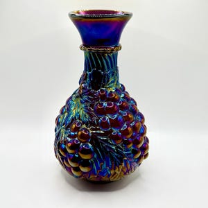 May include: Iridescent glass vase with a bulbous body and a flared rim. The vase is decorated with a grape cluster design and features a vibrant color shift from blue to purple and gold. The vase is approximately 8 inches tall.