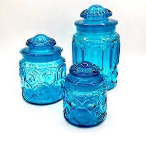 Op de afbeelding: Drie turquoise glazen potten met deksels. De potten hebben verschillende maten en hebben een textuurpatroon van cirkels en sterren. De deksels hebben een decoratief ontwerp. De potten zijn op een witte achtergrond geplaatst.