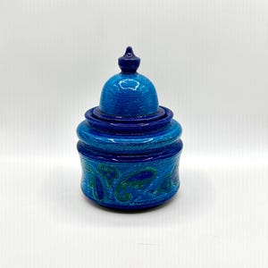 Pode incluir: Um pote cilíndrico de cerâmica com tampa abobadada a condizer, apresentando um remate pontiagudo. O recipiente é vidrado em tons de azul cobalto profundo e turquesa brilhante, decorado com um padrão paisley verde e azul e texturas pontilhadas.
