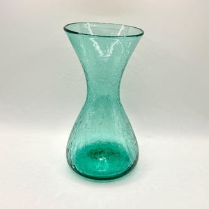 May include: A teal-colored glass vase with a crackle finish. The vase has a wide, flared top that tapers to a narrow neck and then widens again at the base. The glass has a textured appearance, and the vase is empty.