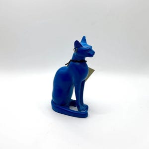 May include: A bright blue figurine of a seated cat with an Egyptian-inspired aesthetic. The cat features a black cord necklace around its neck and a small metallic hoop earring attached to one ear. A small paper tag is visible behind the neck.