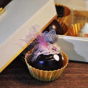 May include: A dark chocolate truffle with a fairy figurine, set in a gold-colored paper cup. The truffle is decorated with pink and purple accents, and is presented in a white box with gold trim. Other chocolates are visible in the background.