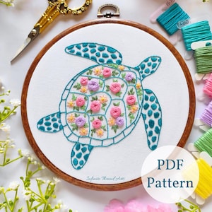 Puede incluir: Patrón bordado de una tortuga marina turquesa con flores rosas, moradas y amarillas en su caparazón. El patrón está en un aro de bordado de madera y rodeado de hilo de bordar.