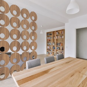 May include: A wooden room divider with a circular cutout pattern. The divider is made of light brown wood and has a modern design. The divider is in a room with a wooden table and chairs.