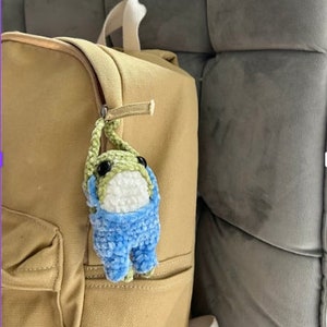 Puede incluir: Un juguete de peluche de rana de crochet verde y azul con ojos blancos colgando de un tirador de cremallera en una mochila de color tostado.