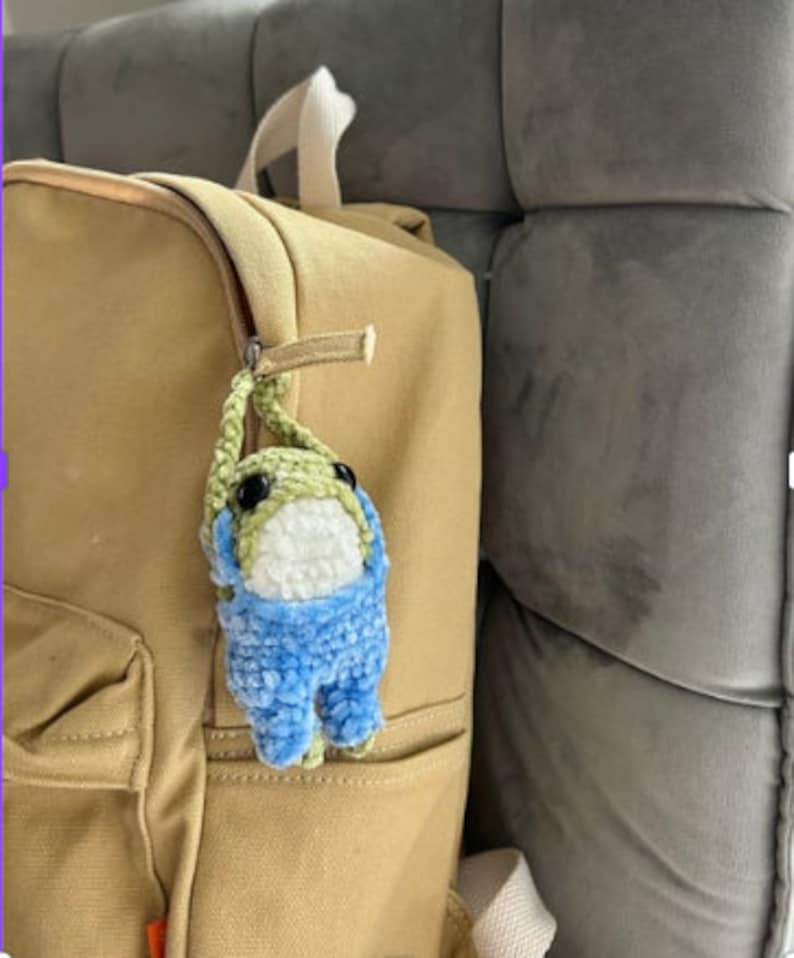 Puede incluir: Un juguete de peluche de rana de crochet verde y azul con ojos blancos colgando de un tirador de cremallera en una mochila de color tostado.