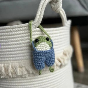 Puede incluir: Un juguete de rana de crochet con un mono azul cuelga de una cesta de cuerda blanca con un borde de flecos.