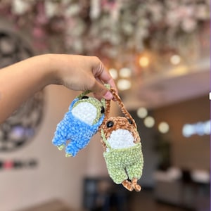 Puede incluir: Dos juguetes de peluche tejidos a crochet colgando de una mano. Uno es azul y el otro es verde con detalles marrones.