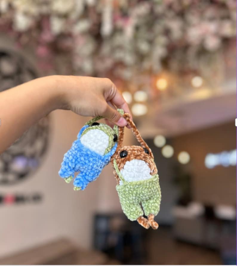 Puede incluir: Dos juguetes de peluche tejidos a crochet colgando de una mano. Uno es azul y el otro es verde con detalles marrones.