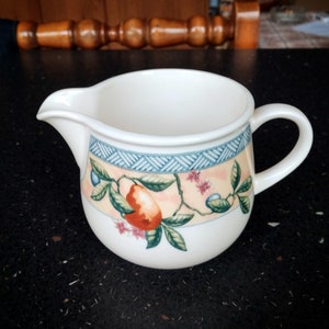 Könnte beinhalten: Weißer Keramik-Sahnegießer mit Henkel und Ausguss. Der Sahnegießer hat ein dekoratives Band mit blauem geometrischen Muster und einem pfirsichfarbenen Blumendesign mit Früchten und Blättern. Der Sahnegießer ist zum Servieren von Sahne oder Milch bestimmt.