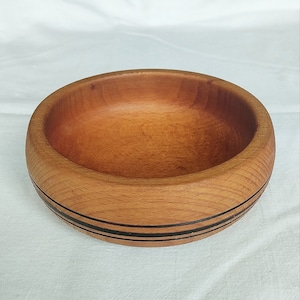 May include: A round wooden bowl with a natural wood grain finish. The bowl has a light brown color with two thin black stripes around the exterior. The bowl is empty and sits on a white surface.