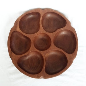 May include: A round, wooden serving tray with seven compartments. The tray is made of a dark brown wood and has a natural, organic shape. The compartments are various sizes and shapes, perfect for serving snacks or appetizers.