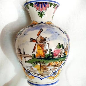 May include: A hand-painted ceramic vase depicting a Dutch windmill scene. The vase has a cream base with a blue rim and floral details. The windmill scene shows a yellow and orange windmill, houses, and a body of water.