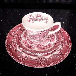 Könnte beinhalten: Ein Vintage-Teeservice mit einem rot-weißen Blumen- und Landschaftsdesign. Das Set beinhaltet eine Teetasse, eine Untertasse und einen kleinen Teller. Die Teetasse hat einen Henkel und ein Blumenmuster am Rand. Die Teller haben eine dekorative Bordüre.
