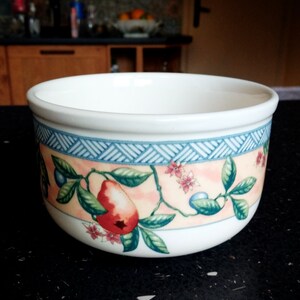 Könnte beinhalten: Weiße Keramikschale mit einem dekorativen Band mit einem blauen geometrischen Muster und einem Design aus roten Birnen, rosa Blüten und grünen Blättern. Die Schale hat eine abgerundete Form und eine glatte, glänzende Oberfläche.