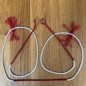 May include: Two white circular cords with red metal knitting needles and red tassels. The needles have a loop at one end and a hook at the other. The tassels are made of red yarn. The items are on a wooden surface.
