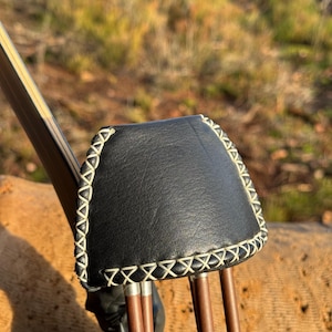 Peut inclure: Carquois de tir à l'arc en cuir noir avec coutures blanches et bord décoratif. Le carquois contient plusieurs flèches en bois avec des pointes métalliques. L'arc est visible en arrière-plan, dans un cadre naturel.