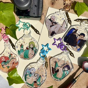 Könnte beinhalten: Eine Sammlung klarer, flaschenförmiger Schlüsselanhänger mit farbenfrohen Illustrationen im Anime-Stil im Inneren. Jeder Schlüsselanhänger hat einen sternförmigen Verschluss und einen stiftförmigen Anhänger. Die Illustrationen zeigen Charaktere in verschiedenen Posen.