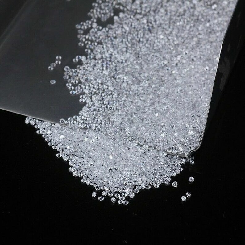 May include: A close-up of a pile of small, clear, round diamonds spilling out of a metal scoop onto a black surface.