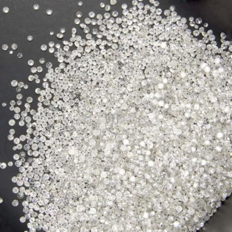May include: A large pile of small, white, round diamonds. The diamonds are loose and scattered on a black surface.