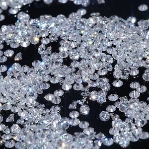 May include: A close-up image of a large quantity of loose, clear, round diamonds scattered on a black surface. The diamonds are reflecting light and sparkle.