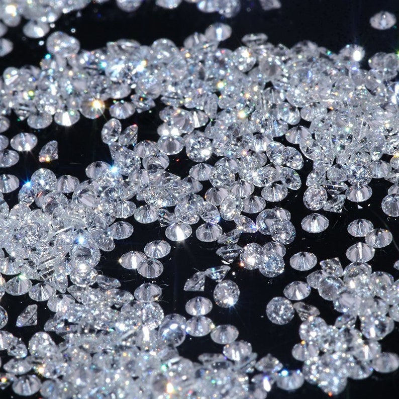 May include: A close-up image of a large quantity of loose, clear, round diamonds scattered on a black surface. The diamonds are reflecting light and sparkle.