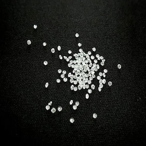 May include: A close-up of a pile of small, clear, round rhinestones scattered on a black surface.