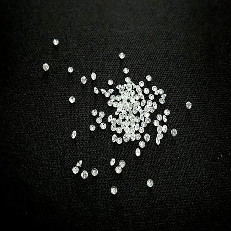 May include: A close-up of a pile of small, clear, round rhinestones scattered on a black surface.