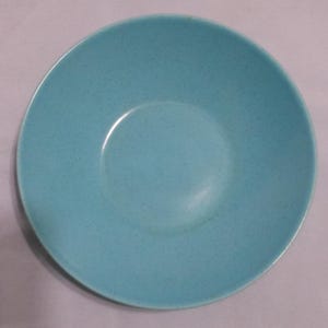 May include: A round, shallow bowl with a smooth, matte finish in a bright turquoise blue color. The surface of the bowl features a subtle, fine speckled pattern throughout, creating a consistent texture across the entire interior basin.