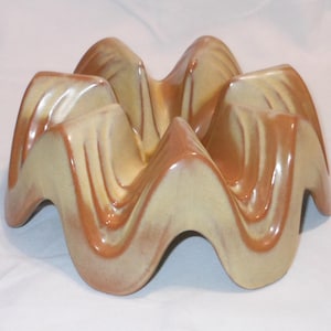 May include: A ceramic bowl with a wavy, undulating rim and a circular base. The piece features a glossy glaze with a two-tone color scheme, blending shades of warm tan and deep terracotta brown in a flowing, rippled pattern across the surface.