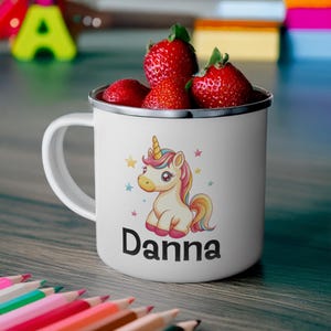 K&ouml;nnte beinhalten: Wei&szlig;e Email Tasse mit einem bunten Comic-Einhorn und dem Namen "Danna" darauf gedruckt. Die Tasse ist mit Erdbeeren gef&uuml;llt.