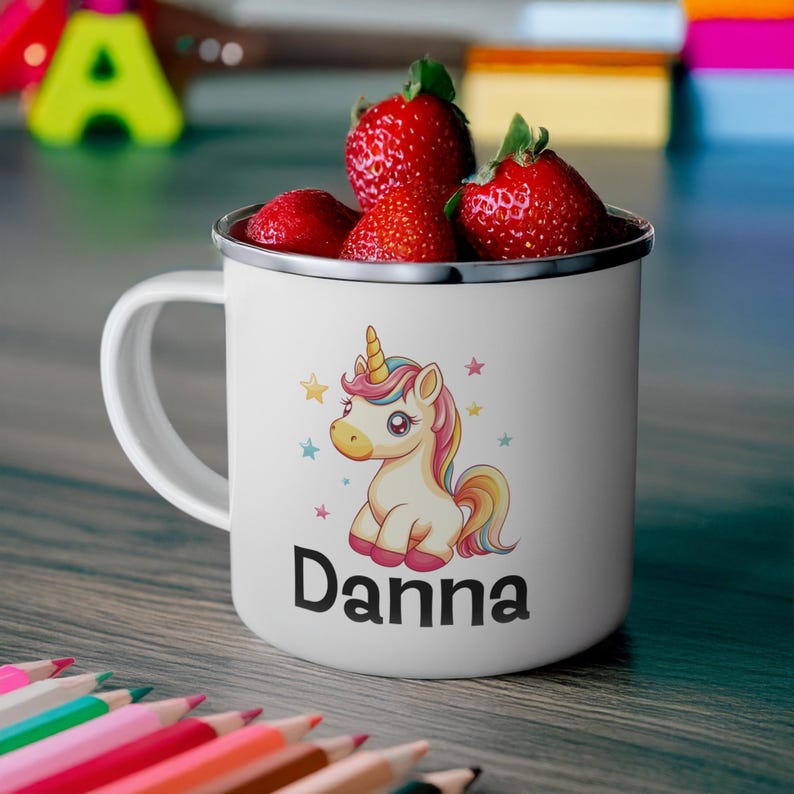 K&ouml;nnte beinhalten: Wei&szlig;e Email Tasse mit einem bunten Comic-Einhorn und dem Namen "Danna" darauf gedruckt. Die Tasse ist mit Erdbeeren gef&uuml;llt.