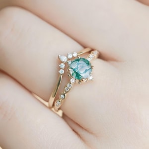 May include: A gold ring set featuring a round, light green gemstone with a unique pattern, surrounded by small, clear accent stones. The set includes a delicate band with additional small stones. The rings are displayed on a hand.