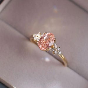May include: A gold ring featuring a large, oval-cut pink gemstone as the centerpiece. The ring is adorned with smaller, clear gemstones clustered around the main stone, creating a sparkling effect. The band is a simple, polished gold.