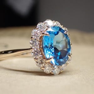 May include: A rose gold ring featuring a large, oval-cut, light blue gemstone surrounded by a halo of smaller, clear gemstones. The ring's band is thin and polished, with a delicate design.