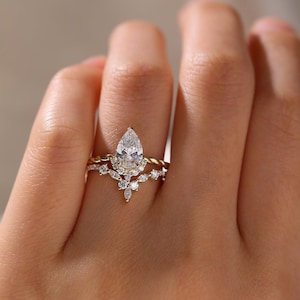 May include: A gold ring set featuring a pear-shaped diamond center stone. The ring is accented with smaller, round diamonds and a twisted band. The set includes a matching band with a floral design.