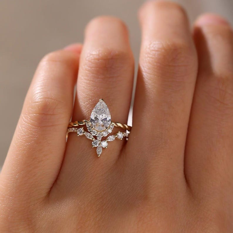 May include: A gold ring set featuring a pear-shaped diamond center stone. The ring is accented with smaller, round diamonds and a twisted band. The set includes a matching band with a floral design.