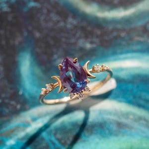 May include: A gold ring featuring a pear-shaped, color-changing alexandrite gemstone. The ring is adorned with crescent moon and small diamond accents. The ring's band is thin and delicate, with a polished finish. The gemstone is a deep purple color.