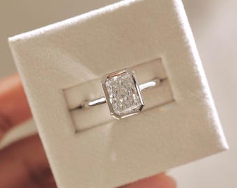 May include: A radiant-cut diamond ring set in a white gold or platinum band. The rectangular diamond is bezel-set, creating a modern and secure design. The ring is displayed in a white jewelry box, highlighting its brilliance.