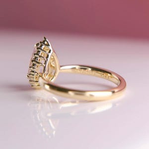 May include: A gold ring with a pear-shaped gemstone. The ring's band and setting are gold. The gemstone is a light pink color and is surrounded by smaller stones. The ring is set against a soft pink background.